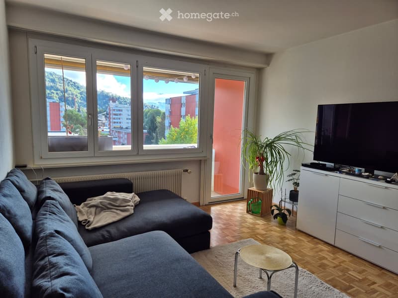 Modern living room with large windows offering a scenic view, a grey sectional sofa, a small round coffee table, a TV on a white cabinet, and several potted plants.
