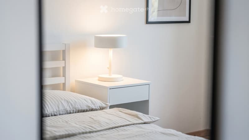 Modern bedroom with a neatly made bed, nightstand, table lamp, and a small shelf on the wall.