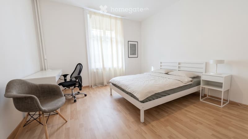 Modern bedroom with a white bed, nightstand, desk, and chair. Light wooden flooring and white walls.