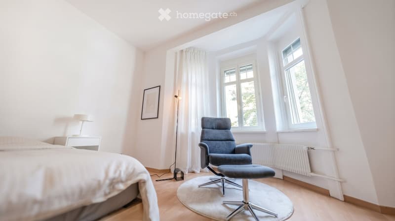 Bright bedroom with wooden flooring, large windows, a bed, a grey armchair, a matching stool, a table lamp, and a picture frame on the wall.