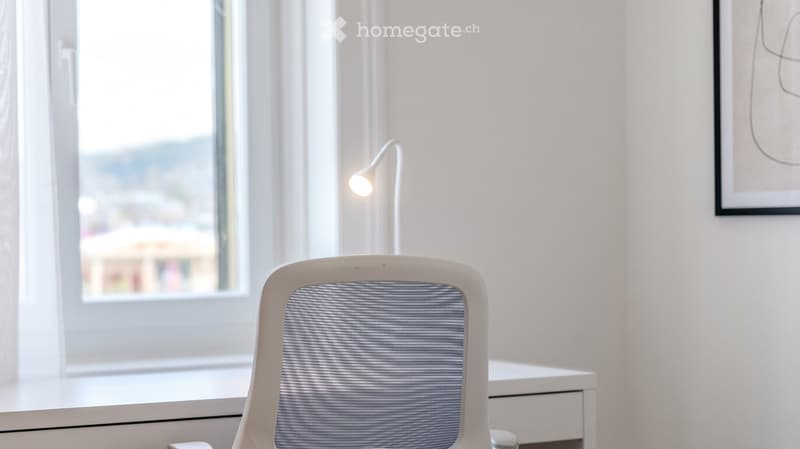 A modern, minimalist room with a white chair, a small desk, a wall-mounted lamp, and a framed picture on the wall. The room has large windows with white curtains, allowing natural light to fill the space.
