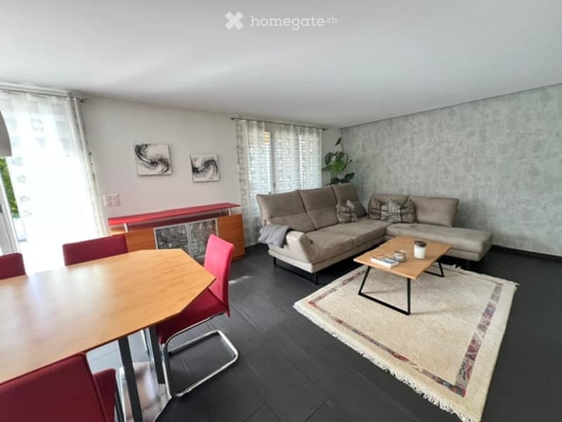 Modern living room with a grey sectional sofa, wooden coffee table, dining table with red chairs, and a patterned rug.