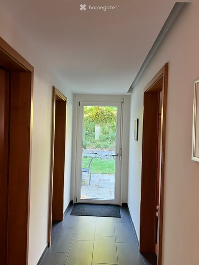 A narrow hallway with dark tiled flooring, white walls, and wooden doors on both sides. A glass door at the end leads to a garden area with a bench visible outside.