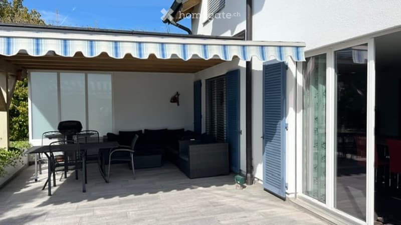 Patio area with a striped awning, outdoor furniture including a sofa, dining table, and chairs, large glass doors leading inside, and blue shutters.