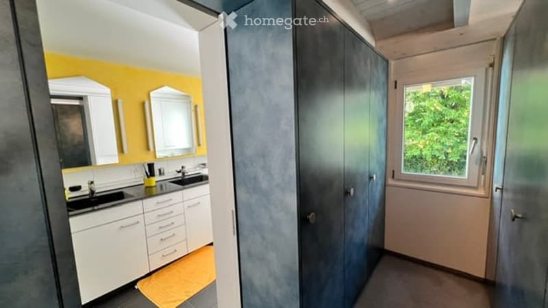 Modern bathroom with double sinks, white cabinets, mirrors, and a large window letting in natural light. The walls are painted in bright yellow and grey, and a yellow towel is placed on the floor.