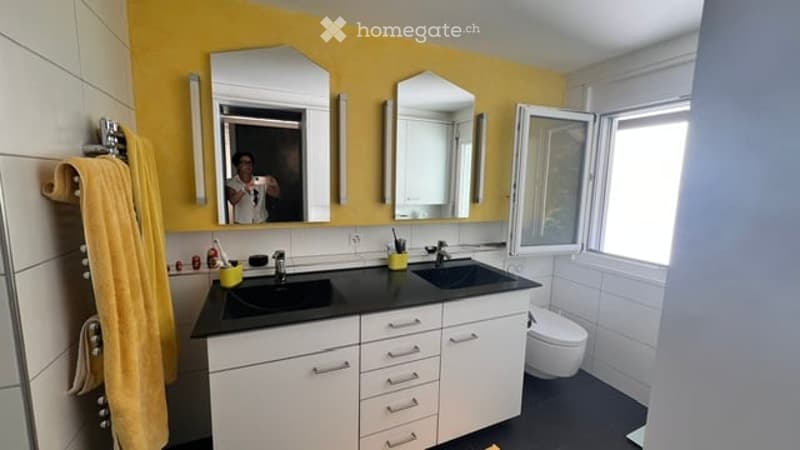 Modern bathroom with double sink vanity, two mirrors, white cabinets, black countertops, and a toilet. Yellow accent wall and white tiles.