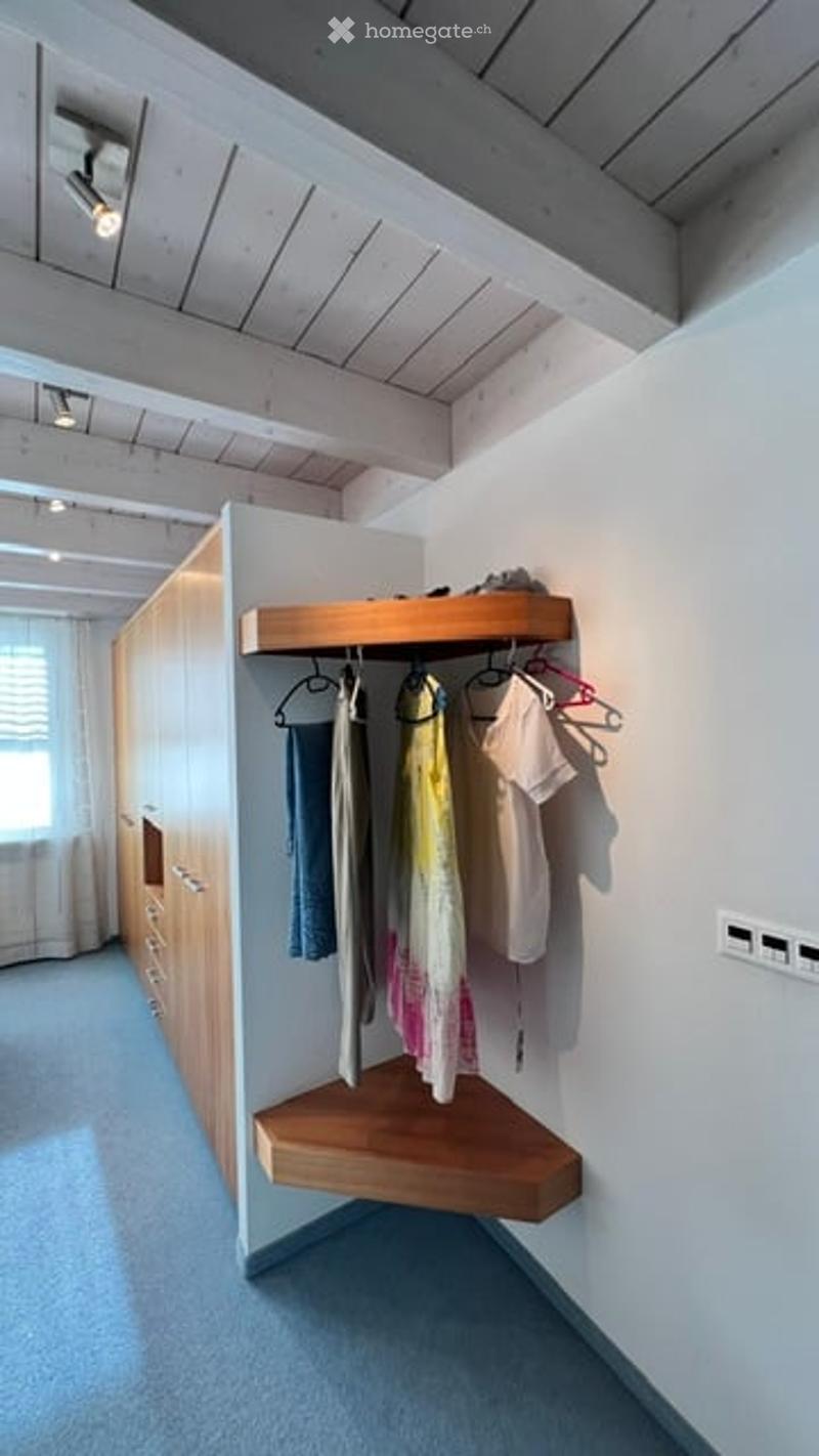 A modern, minimalist walk-in closet with wooden built-in wardrobe and shelves, featuring hanging clothes and storage space.