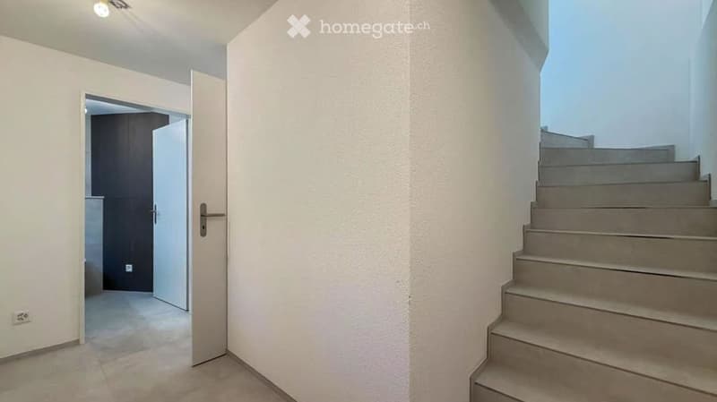 A modern hallway with a staircase leading upwards, white walls, and a door leading to another room.