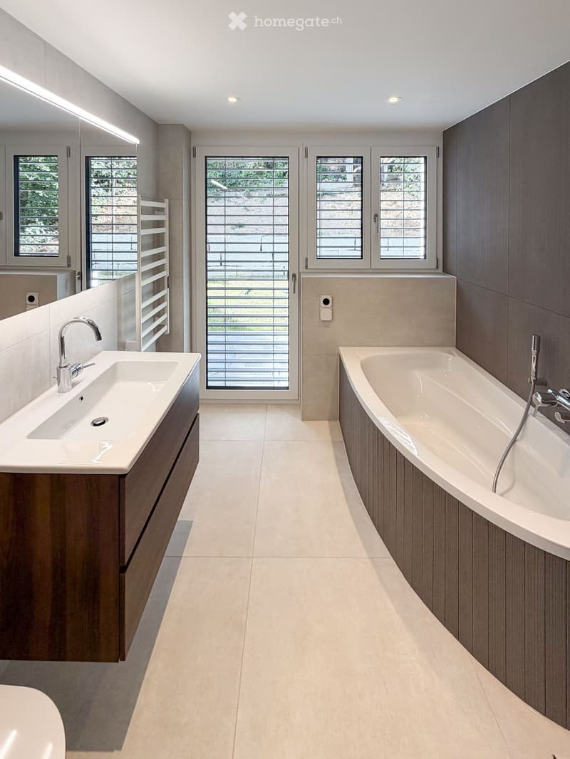 Modern bathroom with a large bathtub, a sink with a modern faucet, and large windows with blinds. The walls are tiled in a light beige color, and the floor is tiled in a light beige color as well.