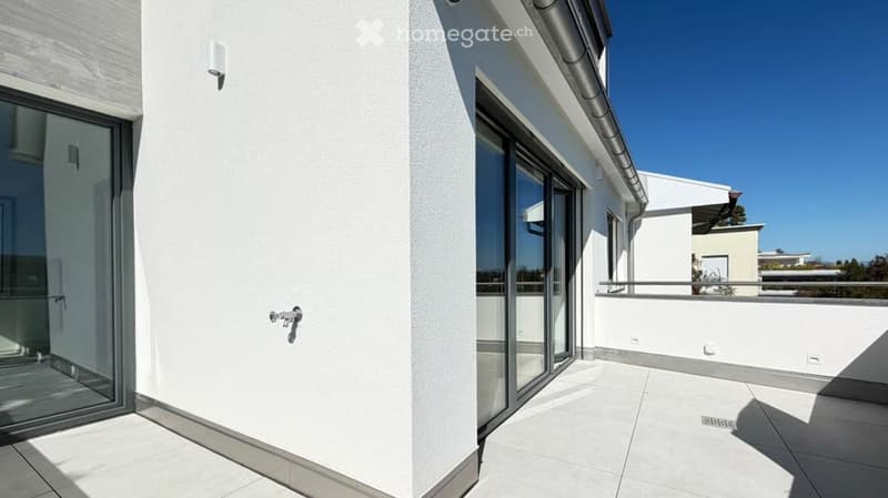 Modern white balcony with large glass doors, tiled floor, and metal railing, overlooking a clear blue sky and neighboring buildings.