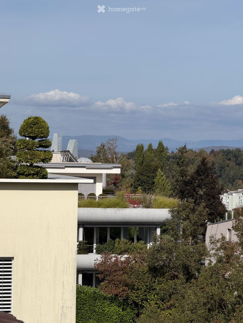 Modern multi-story building with a flat roof, large windows, and surrounding greenery, with distant city skyline and mountains visible.