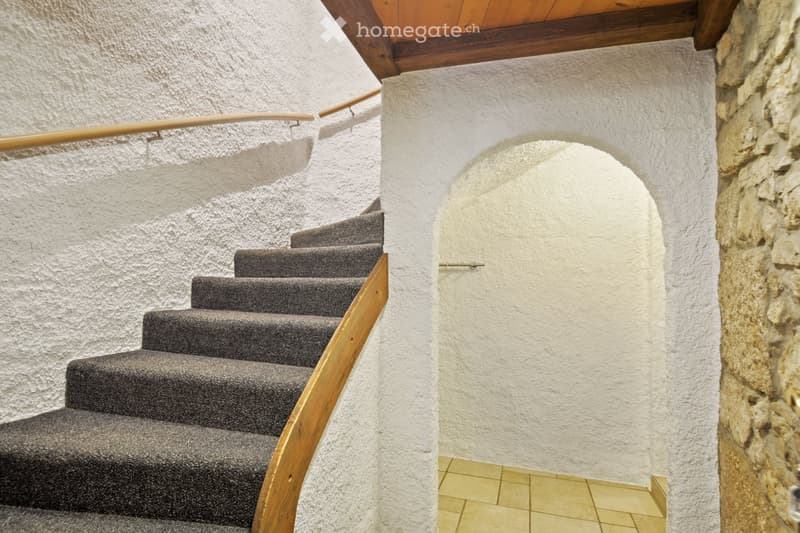 Staircase with wooden handrail and gray carpeted steps, white textured walls, and a stone wall on the right side.