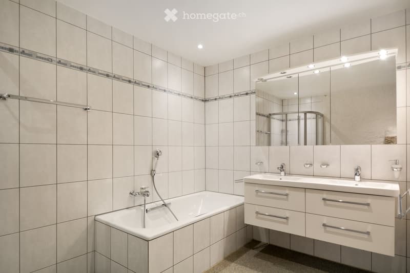 Modern bathroom with a bathtub, double sink, large mirror with integrated lighting, and tiled walls.