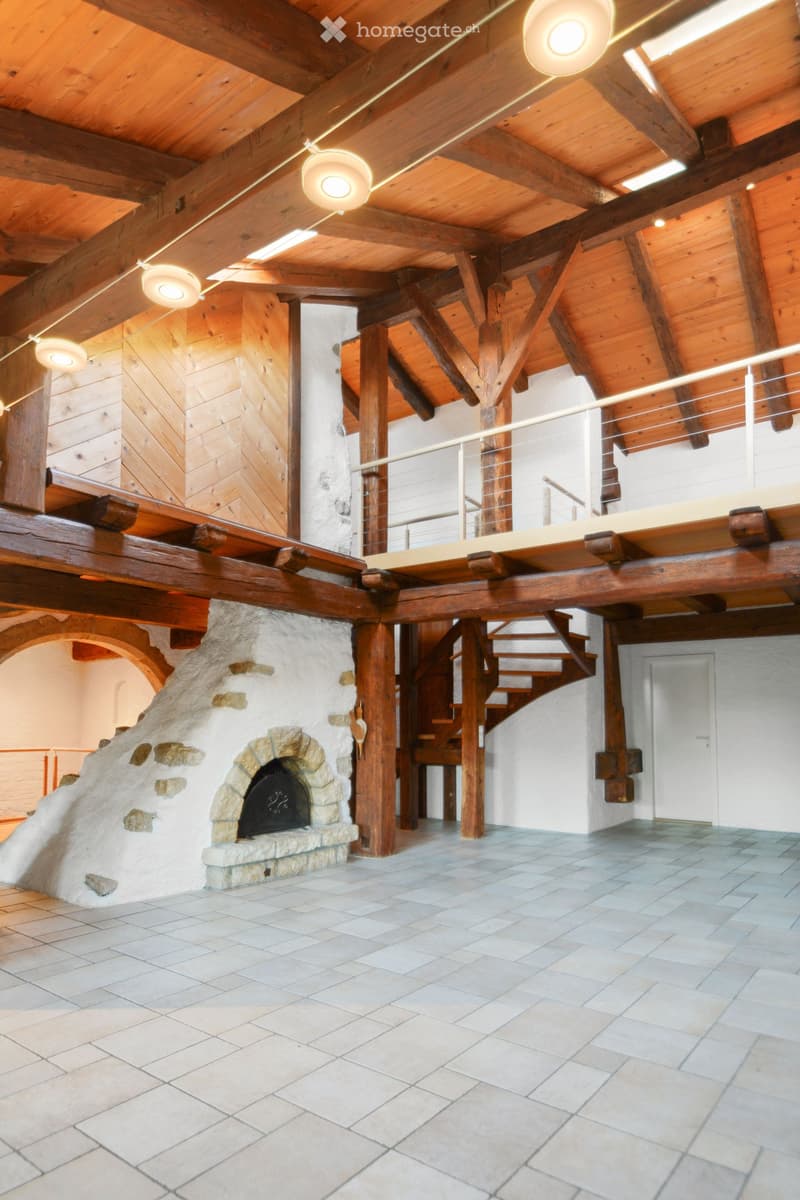 Spacious room with exposed wooden beams, a stone fireplace, and a staircase leading to an upper level.