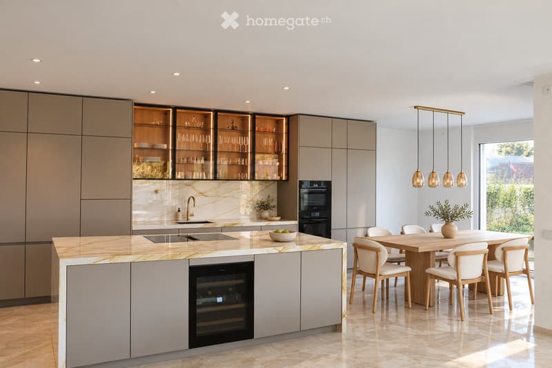 Modern kitchen with light-colored cabinets, marble countertops, built-in oven, wine cooler, and a central island with a sink and pendant lights. A dining table with chairs is positioned near a large window with a view of greenery.