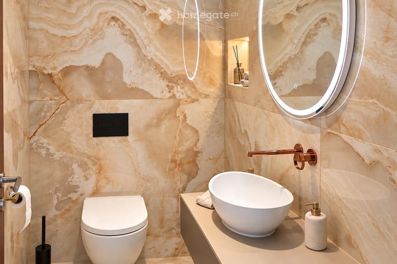 Modern bathroom with marble walls, a white toilet, a sleek sink with a modern faucet, a round illuminated mirror, and a soap dispenser on the counter.
