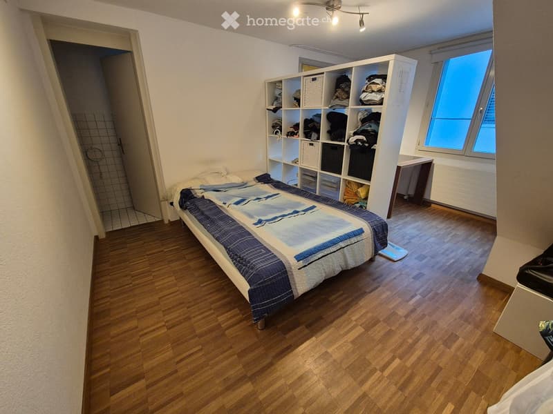 Cozy bedroom with wooden flooring, a neatly made bed, a large storage unit, a small table, and a mirror. The room has large windows and a door leading to a tiled shower area.