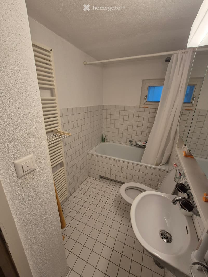 Small bathroom with white tiled walls and floor, featuring a bathtub, toilet, sink, mirror, and a radiator on the left side.