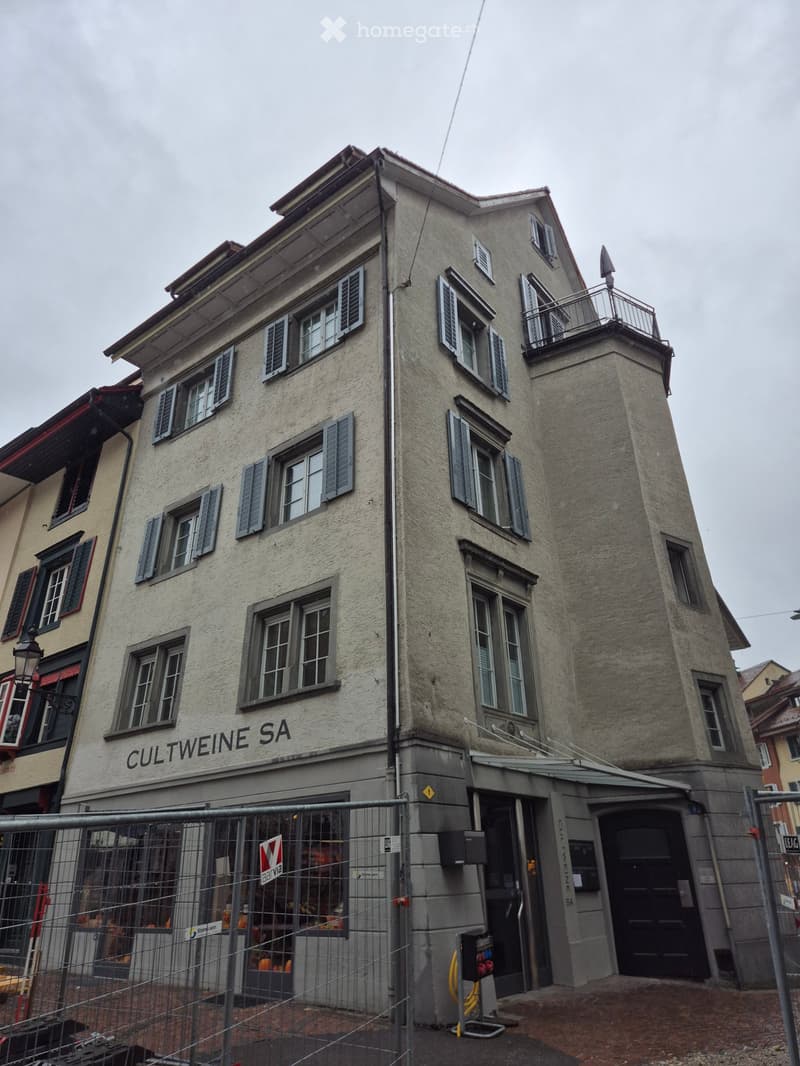Four-story building with light gray facade, multiple windows with shutters, and a balcony on the top floor. The ground floor has a storefront with a sign reading "CULTWEINE SA" and a fenced-off area in front.