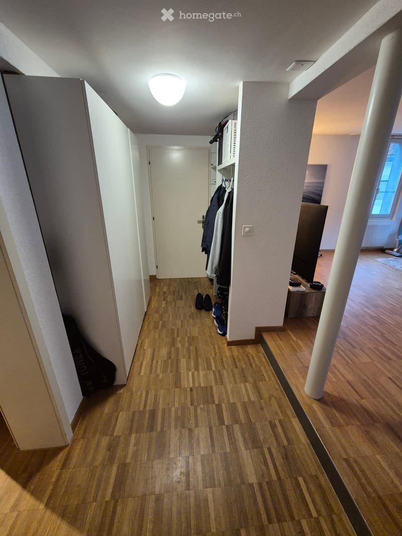 A well-lit hallway with wooden flooring, white walls, and a coat rack holding jackets and shoes. A guitar case is placed against the left wall, and a small table with items sits near the right wall.