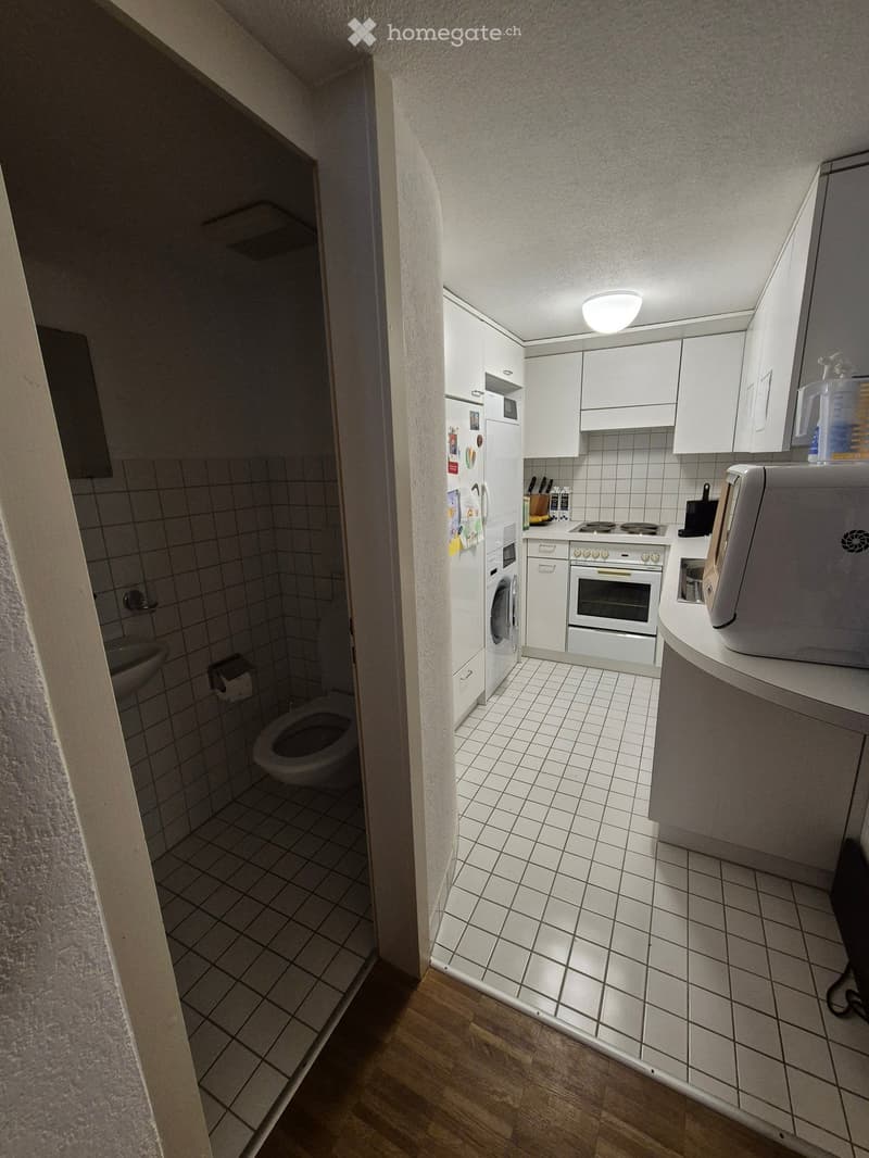 The image shows a compact, well-lit kitchen and bathroom area. The kitchen features white cabinets, a refrigerator, an oven, a microwave, and a toaster. The bathroom includes a toilet, a sink, and a faucet. The floor is tiled, and the walls are also tiled