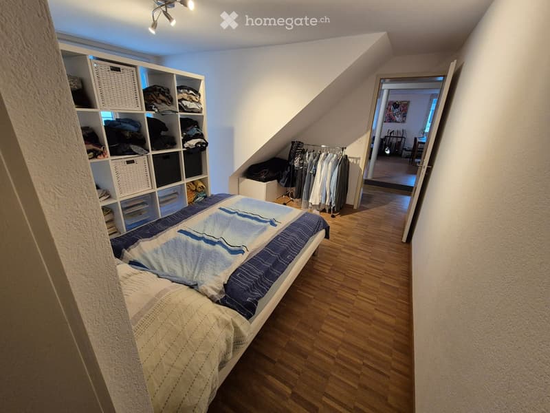 Cozy attic bedroom with wooden flooring, a single bed with blue and white bedding, a built-in shelf unit filled with folded clothes and baskets, and a small clothing rack with hanging garments. The room features exposed wooden beams and a skylight, with a