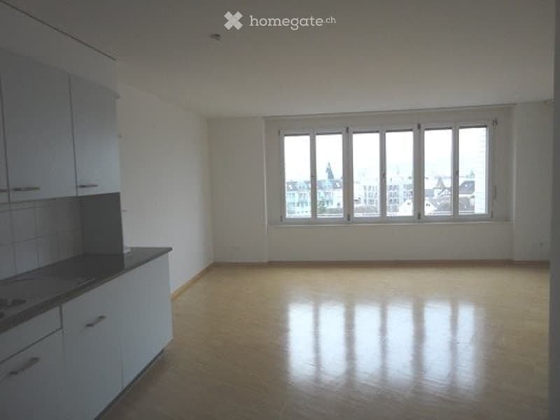Empty room with wooden flooring, white walls, and a fitted kitchen area with cabinets and a countertop. Large windows provide natural light and a view of the city.
