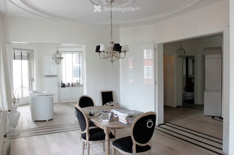 Elegant dining area with a wooden dining table, four chairs, books, a white vase, a white stool, and a chandelier. The room features light-colored walls, wooden flooring, and an open doorway leading to another room.