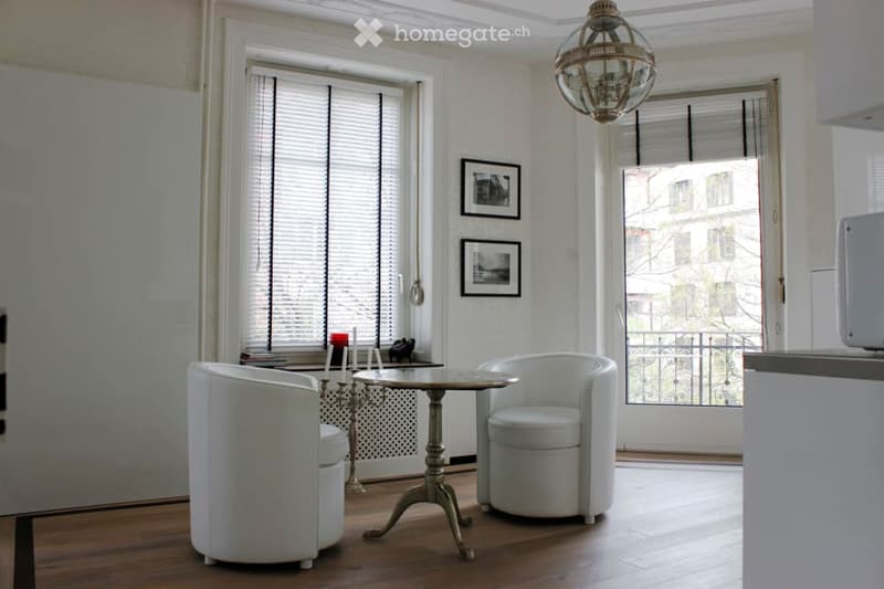 Bright living room with white walls, wooden floor, two white chairs, small table, picture frames, and a chandelier.