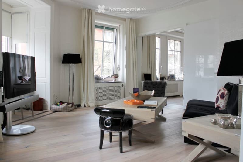 Modern living room with light wooden flooring, large windows with beige curtains, a black sofa with decorative pillows, a sleek coffee table, a modern armchair, a floor lamp, and a flat-screen TV on a stand.
