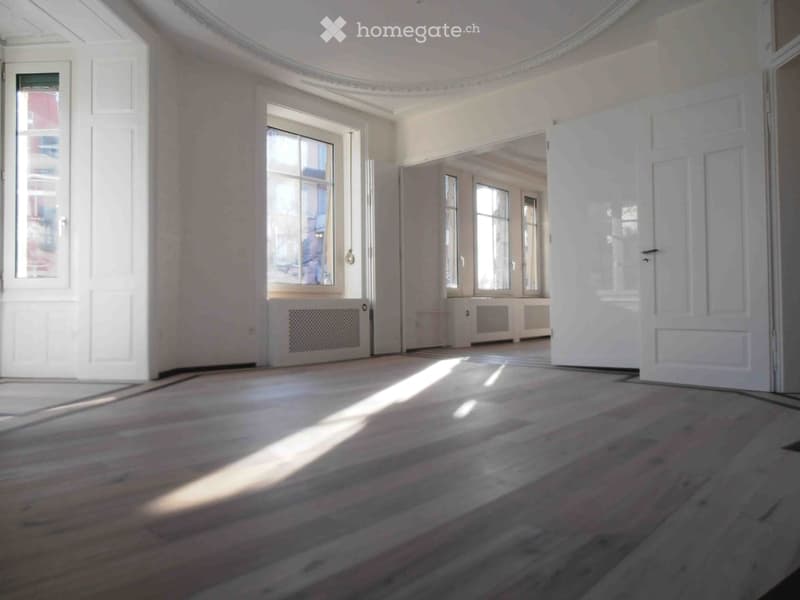Empty room with wooden flooring, large windows, white walls, and radiators. No furniture or equipment present.
