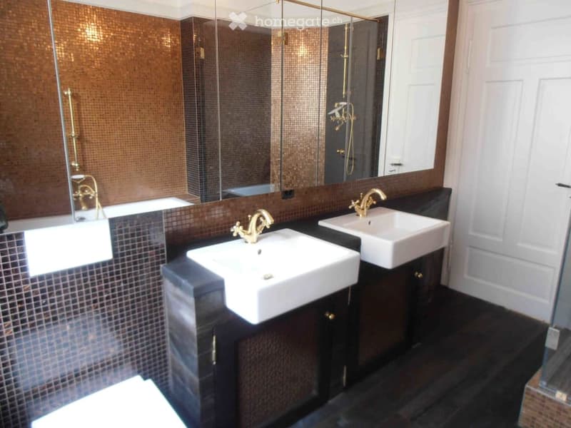 Modern bathroom with two white sinks, gold faucets, a large mirror, and a bathtub, featuring dark tiled walls and floor.