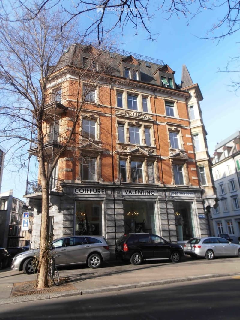 A multi-story brick building with ornate architectural details, featuring storefronts labeled "COIFFURE" and "VALENTINO" with large glass windows and decorative stonework.