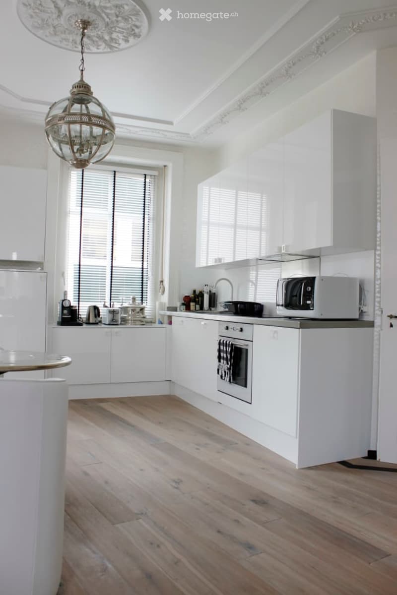 Modern kitchen with white cabinets, wooden flooring, and a hanging chandelier. Features include an oven, microwave, and a coffee maker on the counter.