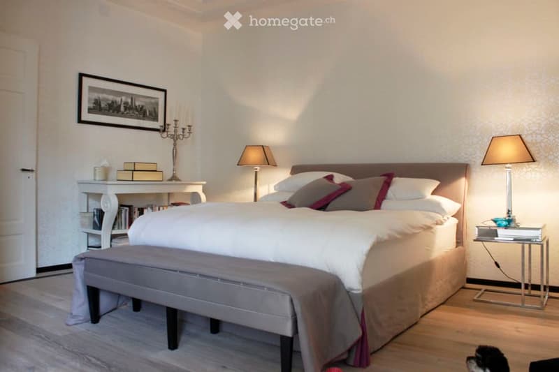 Elegant bedroom with a large bed, nightstands, lamps, a bench at the foot of the bed, and decorative items including a picture frame and candle holder.