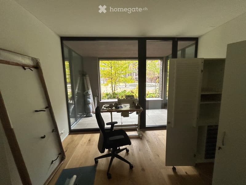 A well-lit room with wooden flooring, a black office chair, a small table with various items, and a large glass door leading to a patio area with outdoor furniture.
