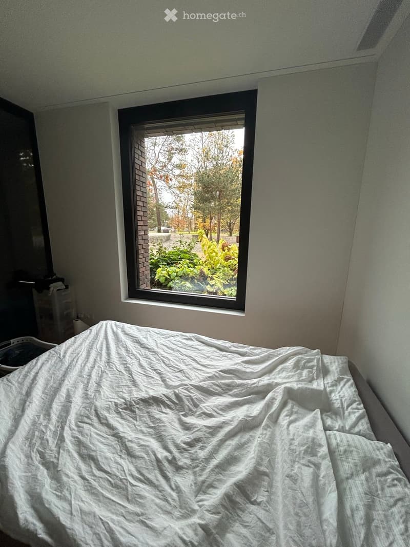 A bedroom with a single bed covered in a white sheet, a window showing a view of greenery and trees, and a small cabinet with items on it.