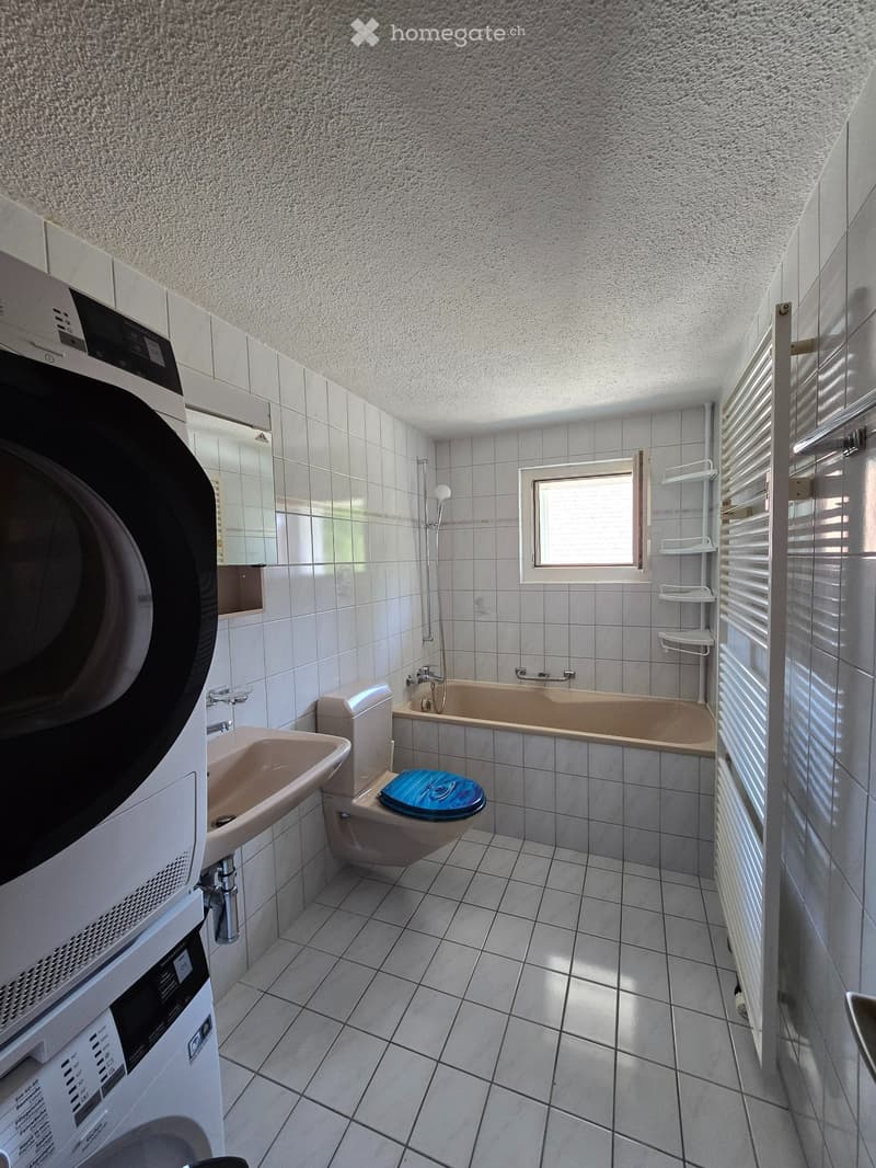 Bathroom with a toilet, sink, bathtub, washing machine, radiator, and a mirror. The room has tiled walls and floor, and a window providing natural light.
