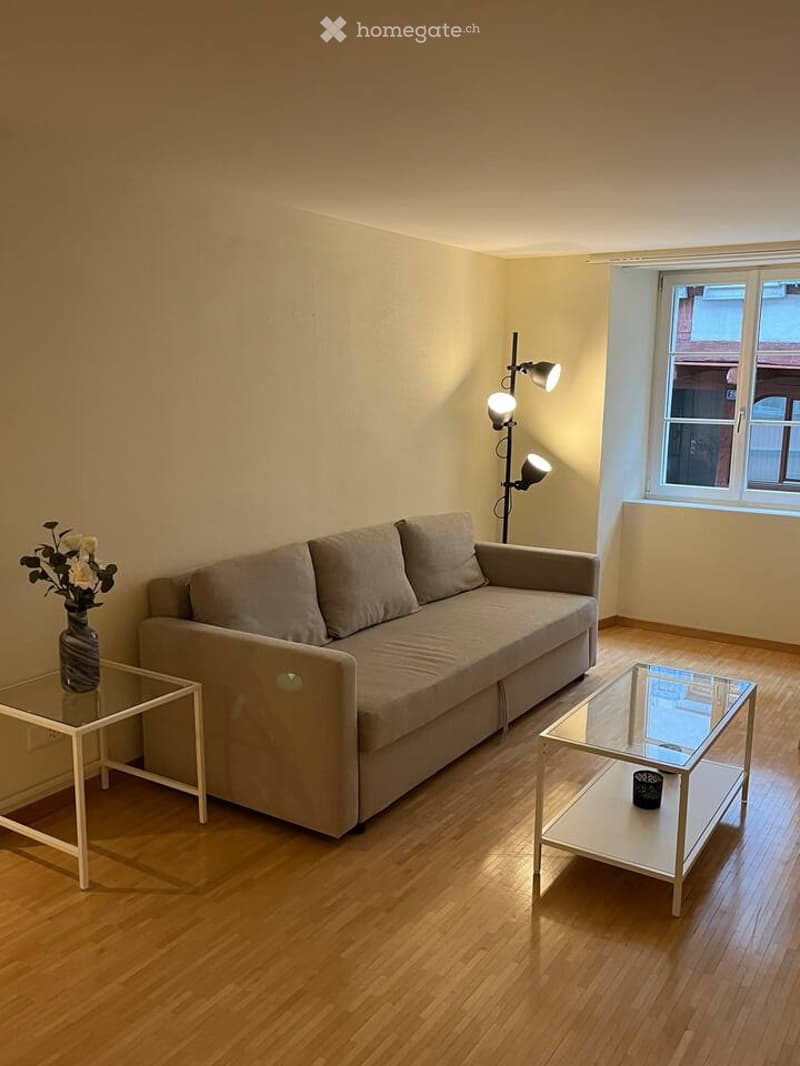 Living room with a grey sofa, two coffee tables, a side table with a vase of flowers, and a floor lamp with multiple spotlights. The room has wooden flooring and large windows.