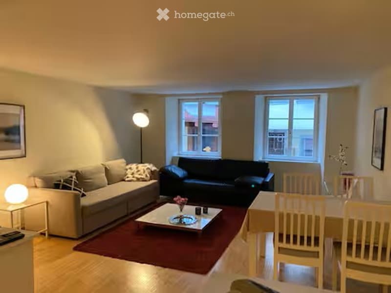 Bright living room with a grey sofa, black armchair, coffee table, dining area with chairs, and a floor lamp. The room features large windows and a warm, inviting atmosphere.