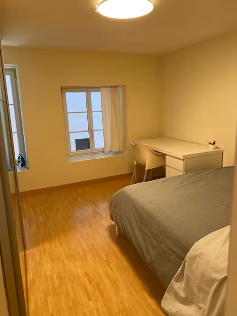 Bright bedroom with wooden flooring, a single bed, a white desk, and a chair. Natural light from two windows with curtains.