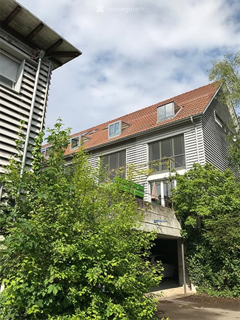 Two-story residential building with a red-tiled roof, white siding, and a balcony on the upper floor. A garage is visible at the front, partially obscured by greenery.