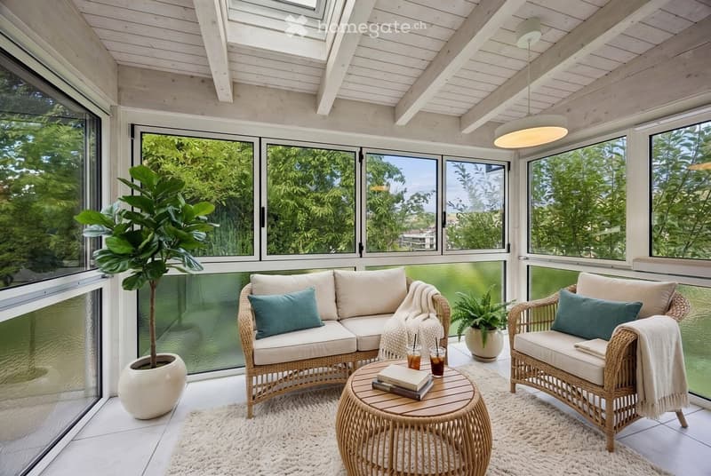 A modern, well-lit balcony with large windows offering a scenic view, furnished with a wicker sofa, matching chair, round coffee table, and potted plants.