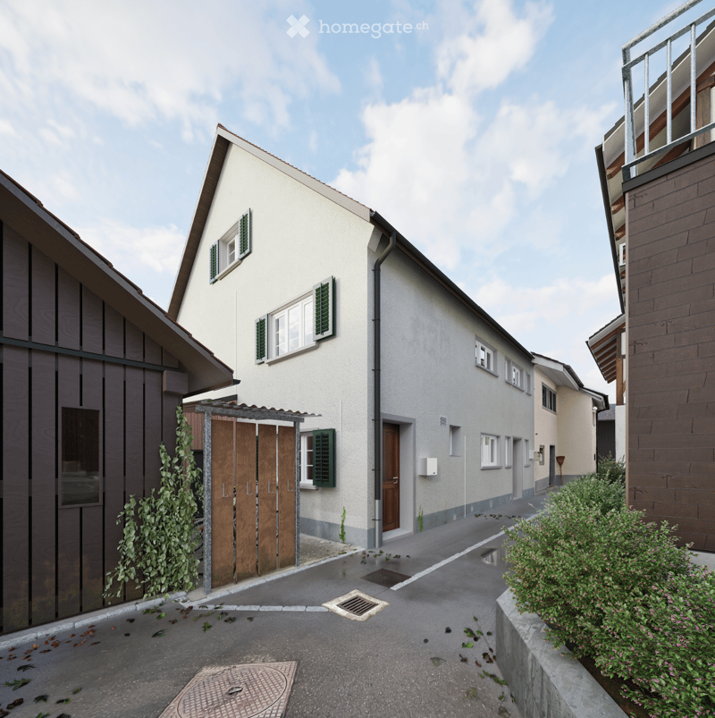A two-story residential building with a white facade, green shutters, and a wooden garage door. The house features a small entrance porch with a wooden door and a small garden area with bushes and flowers.