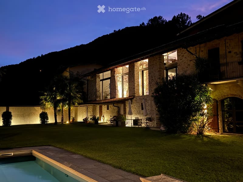 Evening view of a villa's garden with a swimming pool, outdoor seating area, and well-lit pathways.
