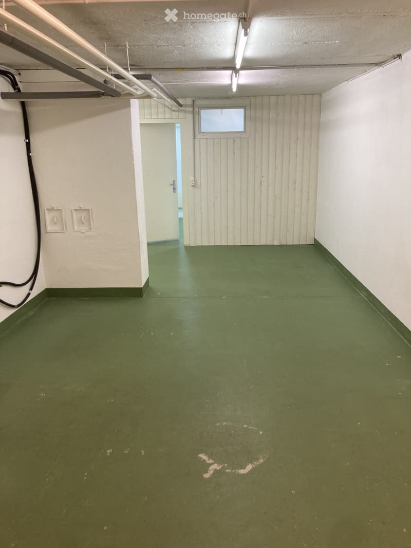 Empty room with green painted floor, white walls, and a single door leading to another space. Exposed pipes and electrical outlets are visible on the walls.