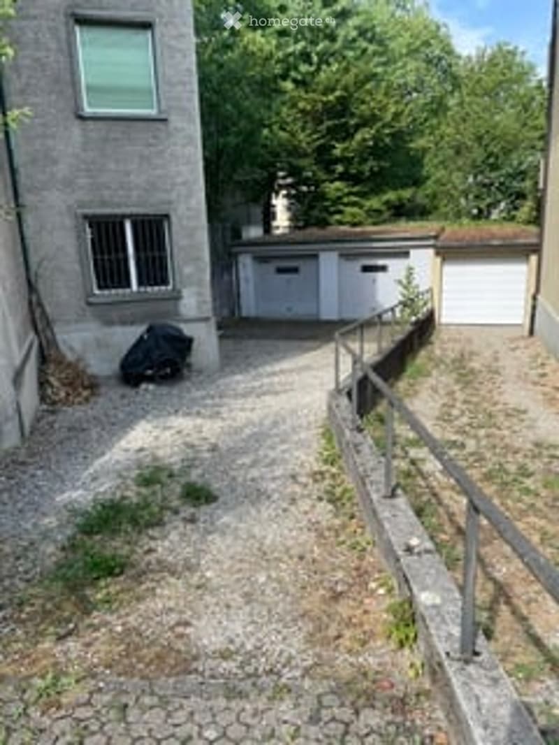 A gravel driveway leads to a building with a garage and a small staircase. The building has a grey facade with windows, and there is a metal fence along the side.