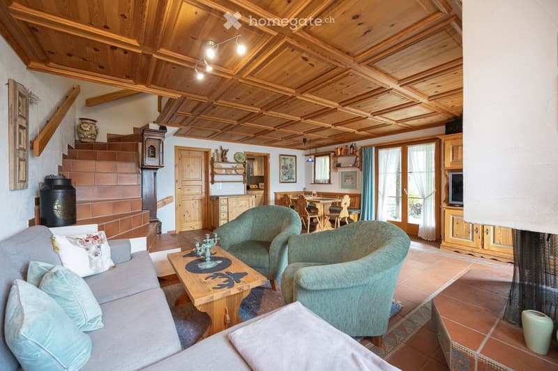 Cozy living room with wooden ceiling beams, tiled floor, grey sofa with pillows, wooden coffee table, armchairs, dining area with table and chairs, television cabinet, and decorative items.