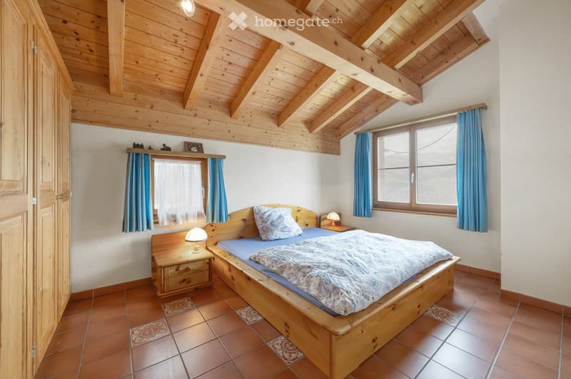 Cozy bedroom with wooden bed, nightstand, lamps, and blue curtains on windows. Exposed wooden ceiling beams and tiled floor.