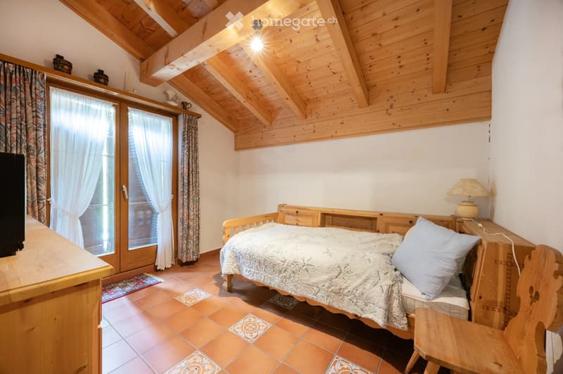 Cozy bedroom with wooden beams, a bed with a patterned quilt, a nightstand with a lamp, a wooden chair, and large windows with curtains.
